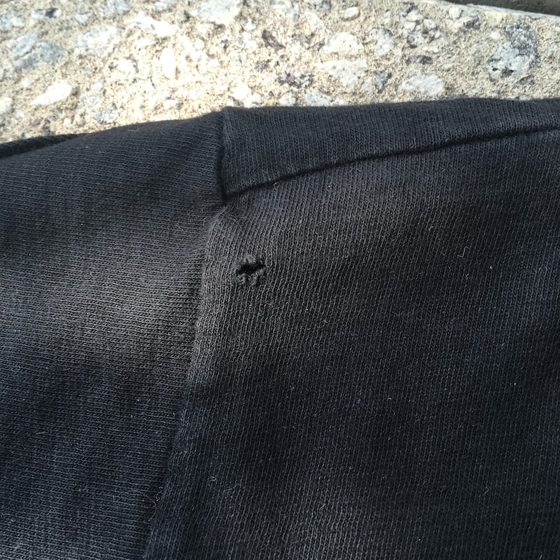 May include: Close-up of a black t-shirt with a small hole near the shoulder seam.