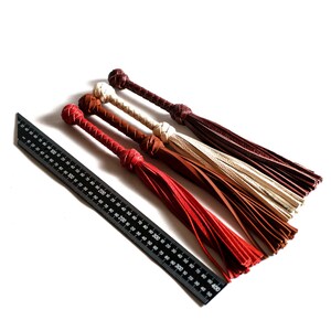 May include: Four leather whips with tassels in red, brown, and cream colors. The whips are laid out on a white surface next to a black ruler.