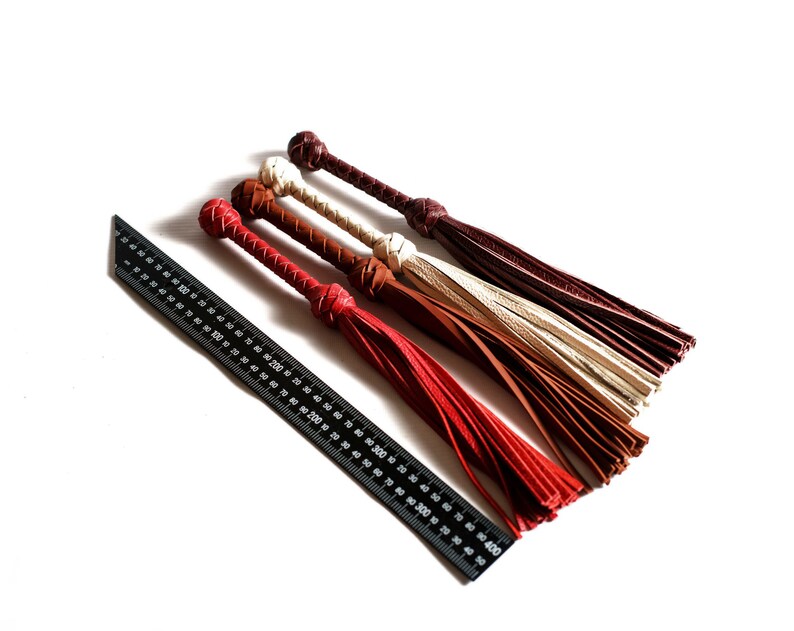 May include: Four leather whips with tassels in red, brown, and cream colors. The whips are laid out on a white surface next to a black ruler.