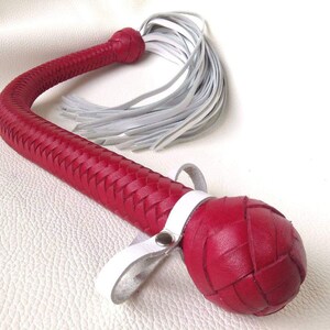 May include: A red leather whip with a braided handle and a white leather tassel. The whip has a white leather strap with a metal snap.