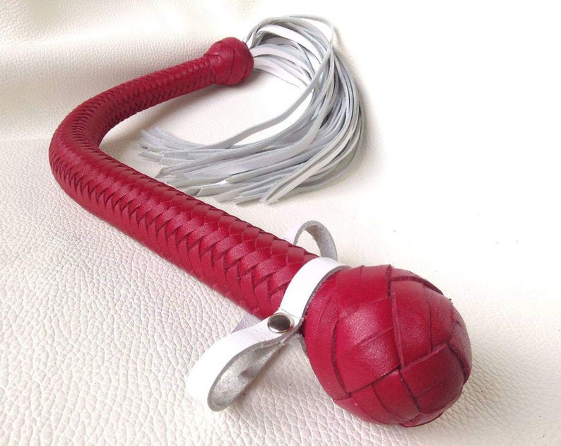 May include: A red leather whip with a braided handle and a white leather tassel. The whip has a white leather strap with a metal snap.