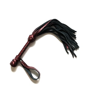 May include: A red leather whip with a black tassel. The whip has a braided handle and a loop for holding.