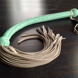 May include: A light green leather whip with a braided handle and a long, thick fringe of light brown leather.