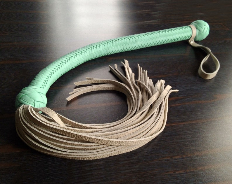 May include: A light green leather whip with a braided handle and a long, thick fringe of light brown leather.