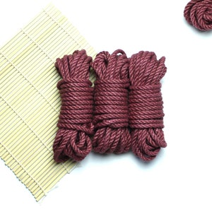 May include: Three spools of thick, maroon-colored rope are arranged on a bamboo mat. The rope is tightly wound and has a slightly textured surface.