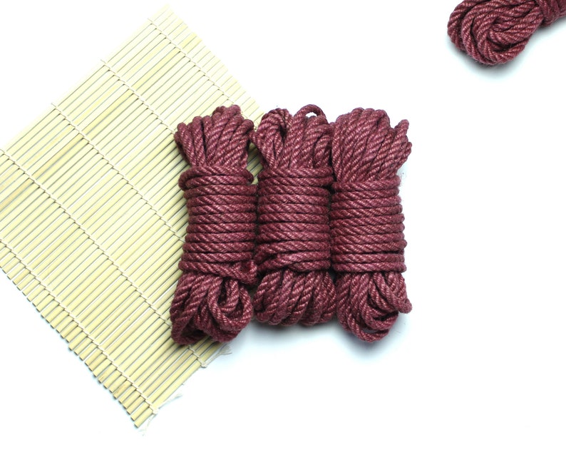May include: Three spools of thick, maroon-colored rope are arranged on a bamboo mat. The rope is tightly wound and has a slightly textured surface.