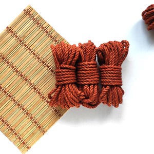 May include: Three spools of thick, rust-colored rope are stacked on a bamboo mat. The rope is coiled tightly and has a rough texture.
