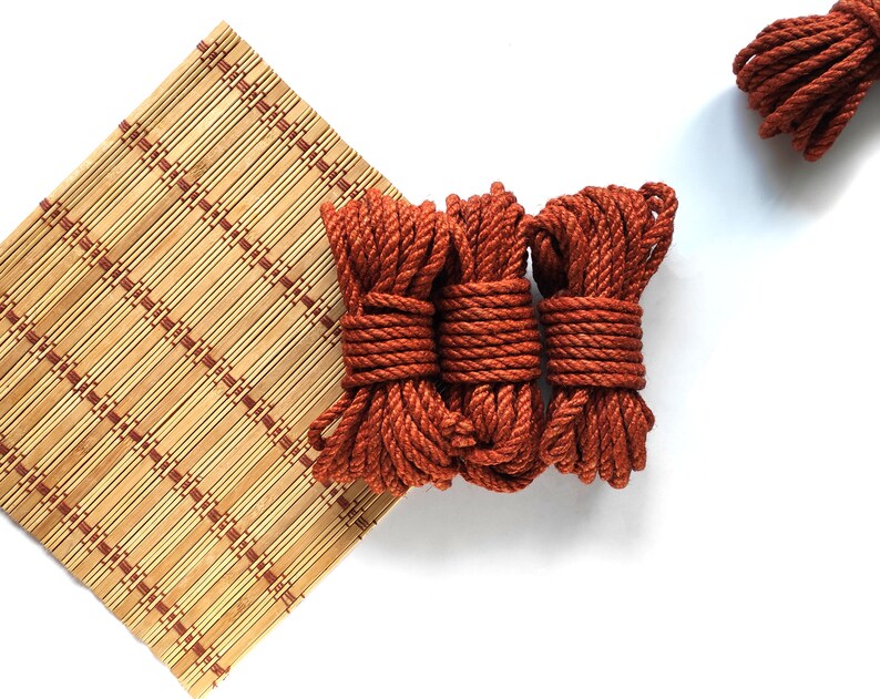 May include: Three spools of thick, rust-colored rope are stacked on a bamboo mat. The rope is coiled tightly and has a rough texture.