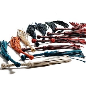 May include: A collection of leather whips with various colored tassels, including teal, orange, white, and burgundy. The whips are arranged in a fan shape on a white background.