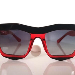Könnte beinhalten: Geometrische Sonnenbrille mit schwarzem oberem Rahmen und rotem unterem Rahmen. Die Gläser sind grau verlaufend. Die Sonnenbrille hat ein modernes, eckiges Design, passend für modebewusste Menschen.