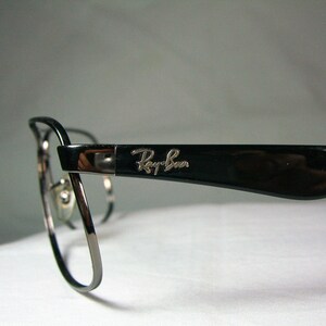 K&ouml;nnte beinhalten: Schwarze Ray-Ban Brille mit silbernem Logo an der Seite. Die Brille hat eine abgerundete, rechteckige Form.