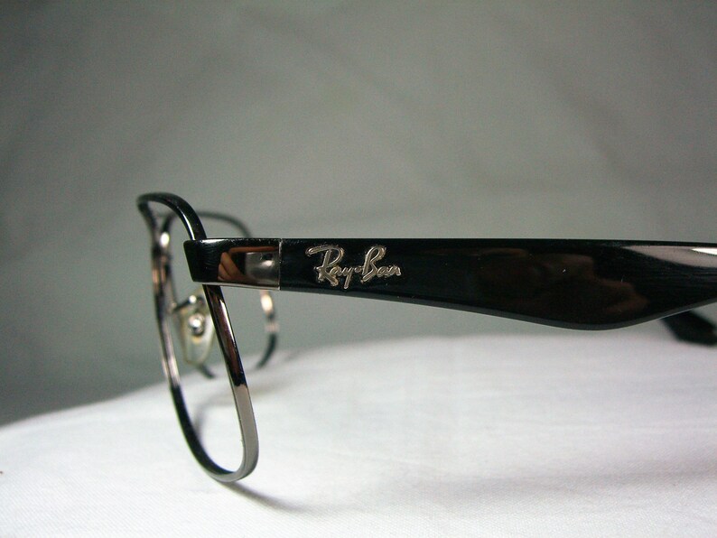 K&ouml;nnte beinhalten: Schwarze Ray-Ban Brille mit silbernem Logo an der Seite. Die Brille hat eine abgerundete, rechteckige Form.