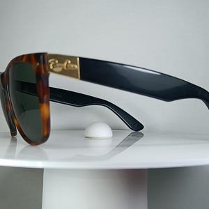 May include: Sunglasses with a tortoiseshell frame and dark lenses. The temples are black with a gold accent. The sunglasses are resting on a white surface.