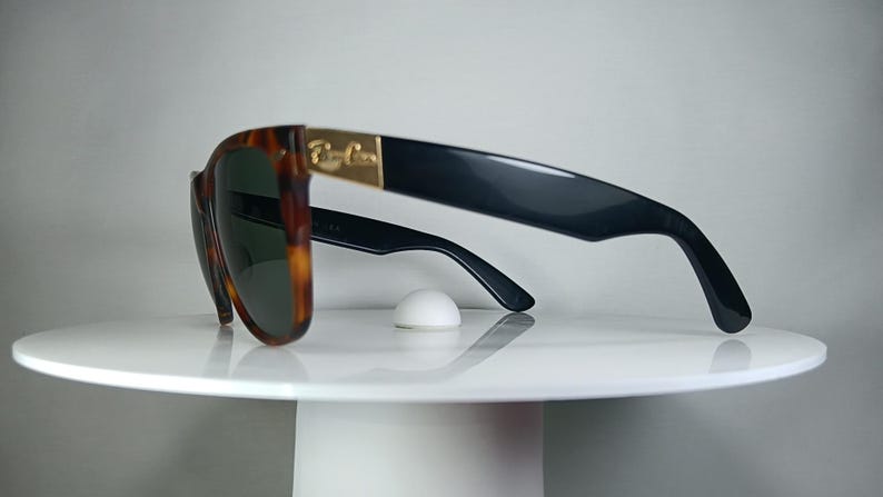 May include: Sunglasses with a tortoiseshell frame and dark lenses. The temples are black with a gold accent. The sunglasses are resting on a white surface.