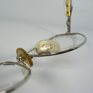 May include: Silver-toned glasses with a gold accent on the arm. The glasses have a clear lens and a small, clear, oval-shaped piece on the frame with the letter "G" embossed on it.