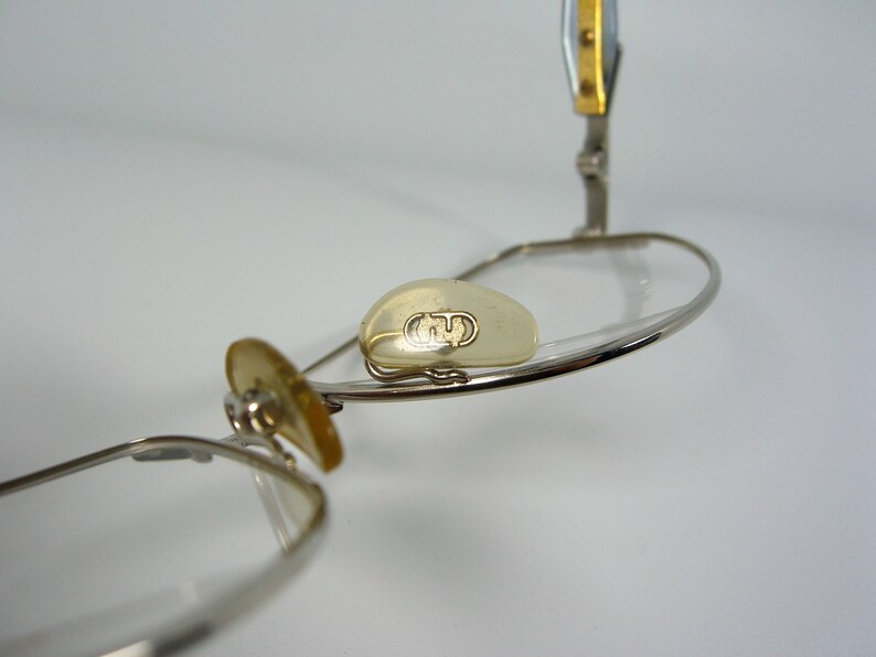 May include: Silver-toned glasses with a gold accent on the arm. The glasses have a clear lens and a small, clear, oval-shaped piece on the frame with the letter "G" embossed on it.