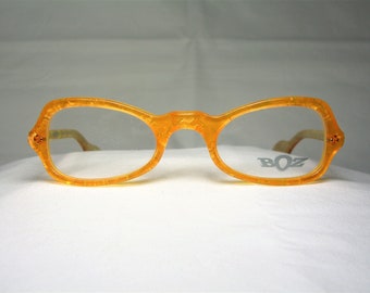Puede incluir: Gafas de plástico de color naranja con lentes transparentes. El nombre de la marca "BOZ" está impreso en la lente derecha.