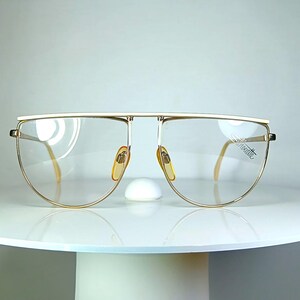 Könnte beinhalten: Goldrandbrille mit einem einzigartigen, übergroßen Design. Die Brille hat einen geraden oberen Bügel und gebogene Gläser, mit einem kleinen Nasensteg und hellen Nasenpads. Die Gläser sind klar, und der Rahmen hat eine Vintage-Ästhetik.