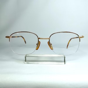 Può includere: Occhiali a mezza montatura color oro con lenti trasparenti. Le montature hanno un design classico con una sottile montatura in metallo dorato e aste con motivo tartarugato. Gli occhiali sono esposti su un supporto in acrilico trasparente.