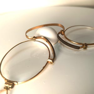 Puede incluir: Tres gafas antiguas doradas con lentes transparentes dispuestas sobre una superficie blanca. Un par está abierto, mientras que los otros están cerrados. Las monturas tienen un diseño delicado y vintage, lo que sugiere un artículo histórico o de colección.