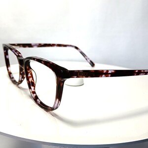 Peut inclure: Lunettes rectangulaires avec un motif tachet&eacute; marron fonc&eacute; et blanc. Les montures ont un bord l&eacute;g&egrave;rement arrondi. Les branches sont &eacute;galement &agrave; motifs. Les lunettes sont pos&eacute;es sur une surface blanche.