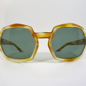 Puede incluir: Gafas de sol vintage de gran tamaño con montura hexagonal. La montura es una degradación de amarillo y naranja, con lentes verde oscuro. Las patillas son de color marrón claro. Fondo blanco.