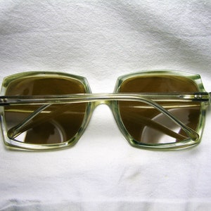 Może przedstawiać: Okulary przeciwsłoneczne z przezroczystego plastiku z brązowymi szkłami. Okulary mają kwadratowy kształt i leżą na białym tle.