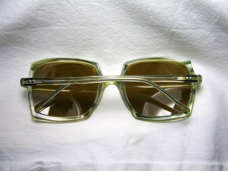 Może przedstawiać: Okulary przeciwsłoneczne z przezroczystego plastiku z brązowymi szkłami. Okulary mają kwadratowy kształt i leżą na białym tle.