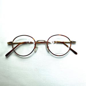 May include: Round eyeglasses with a tortoiseshell pattern on the rims and temples. The frames are gold-toned, and the lenses are clear. The glasses have a classic, vintage style and are set against a white background.