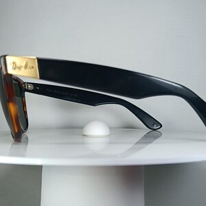 May include: Black sunglasses with a tortoiseshell frame and dark lenses. The sunglasses have a gold accent on the top of the frame and are resting on a white, round surface. The sunglasses are angled to the left.