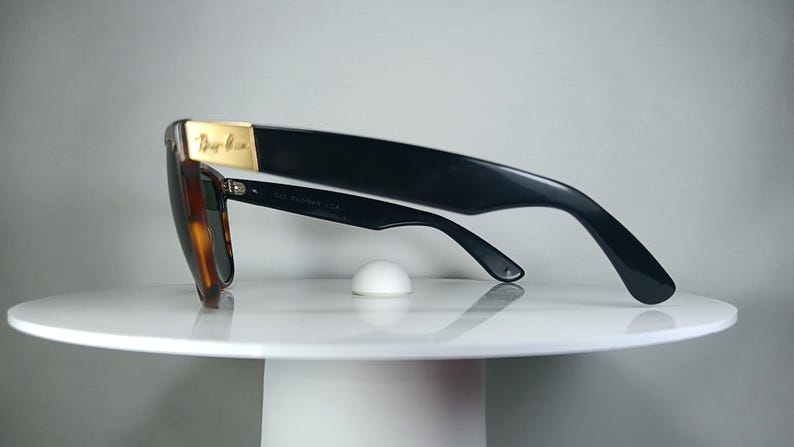 May include: Black sunglasses with a tortoiseshell frame and dark lenses. The sunglasses have a gold accent on the top of the frame and are resting on a white, round surface. The sunglasses are angled to the left.