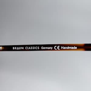 May include: Close-up of the arm of a pair of eyeglasses. The frame is a tortoiseshell pattern. The text "BRAUN CLASSICS Germany CE Handmade" is printed in white on the arm of the glasses.