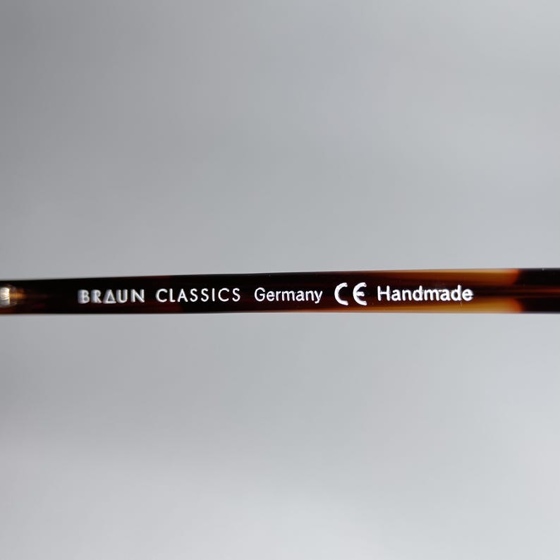 May include: Close-up of the arm of a pair of eyeglasses. The frame is a tortoiseshell pattern. The text "BRAUN CLASSICS Germany CE Handmade" is printed in white on the arm of the glasses.