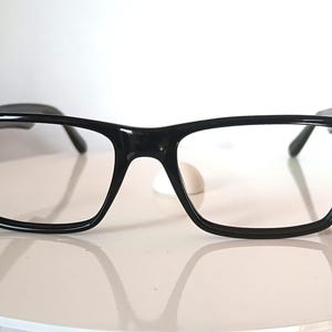 Puede incluir: Gafas negras con montura rectangular. Las gafas tienen una montura gruesa y brillante y lentes transparentes. Las patillas son finas y se extienden hacia atrás hasta la oreja. Las gafas están sobre una superficie blanca.