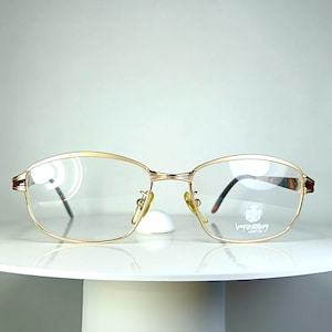 May include: Gold-rimmed eyeglasses with clear lenses. The oval frames have a subtle design and are set on a white surface. The temples are dark brown. The lenses have a logo on the right lens.