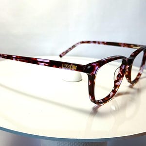 Peut inclure: Lunettes Roxy avec un motif &eacute;caille de tortue dans les tons bordeaux, violet et cr&egrave;me. Les montures rectangulaires ont une forme subtile en &oelig;il de chat. Le logo Roxy est visible sur la branche. Les lunettes sont pr&eacute;sent&eacute;es sur une surface blanche.