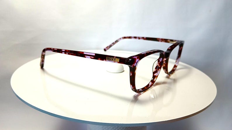 Peut inclure: Lunettes Roxy avec un motif &eacute;caille de tortue dans les tons bordeaux, violet et cr&egrave;me. Les montures rectangulaires ont une forme subtile en &oelig;il de chat. Le logo Roxy est visible sur la branche. Les lunettes sont pr&eacute;sent&eacute;es sur une surface blanche.