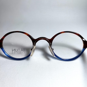 May include: Round eyeglasses with a brown and blue frame. The top of the frame has a tortoiseshell pattern, while the bottom is blue. The lenses are clear, and the nose pads are transparent. The brand name "BRAUN CLASSICS" is printed on the lens.