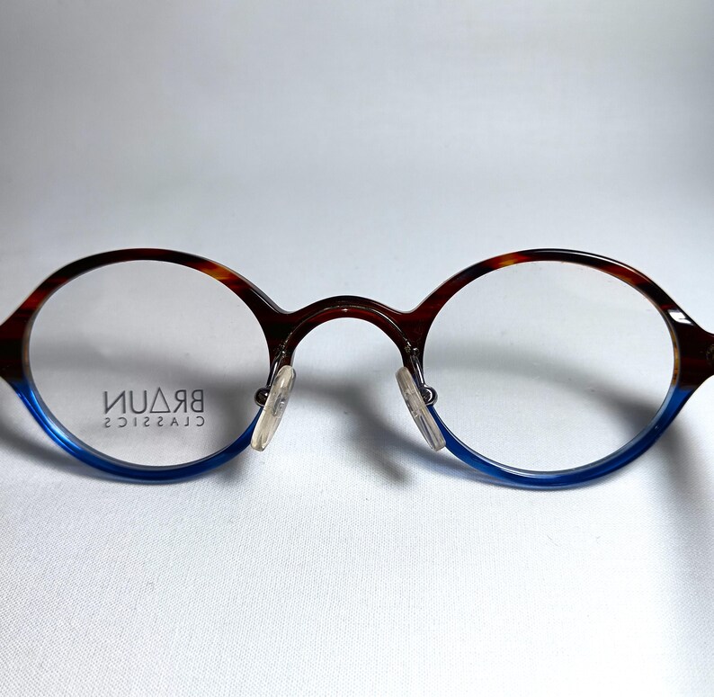 May include: Round eyeglasses with a brown and blue frame. The top of the frame has a tortoiseshell pattern, while the bottom is blue. The lenses are clear, and the nose pads are transparent. The brand name "BRAUN CLASSICS" is printed on the lens.