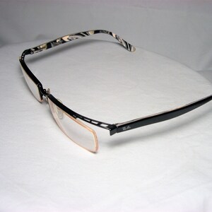 May include: Black and white patterned eyeglasses with a black frame and clear lenses. The glasses are folded in half and the Ray-Ban logo is visible on the right arm.