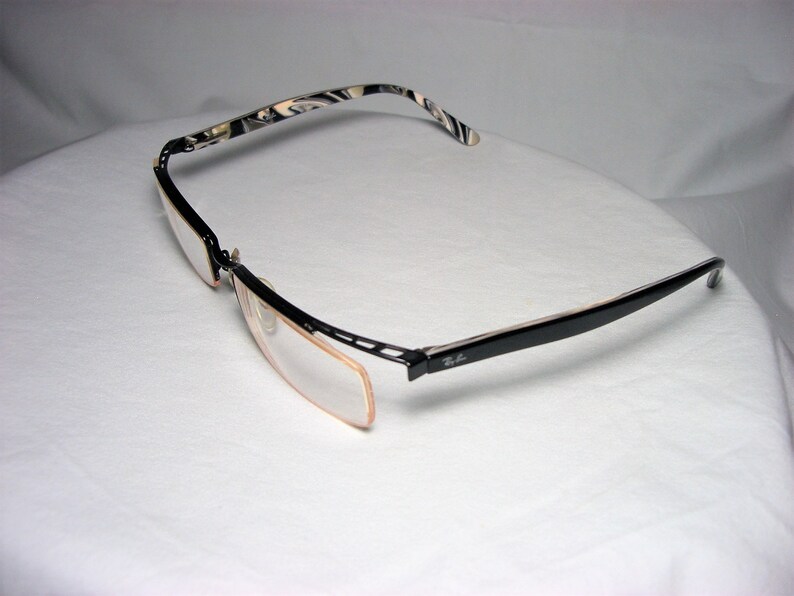 May include: Black and white patterned eyeglasses with a black frame and clear lenses. The glasses are folded in half and the Ray-Ban logo is visible on the right arm.