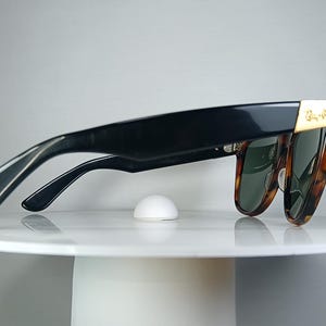 May include: Black sunglasses with a tortoiseshell pattern on the frames and green lenses. The sunglasses have a gold accent on the temples and are resting on a white surface.