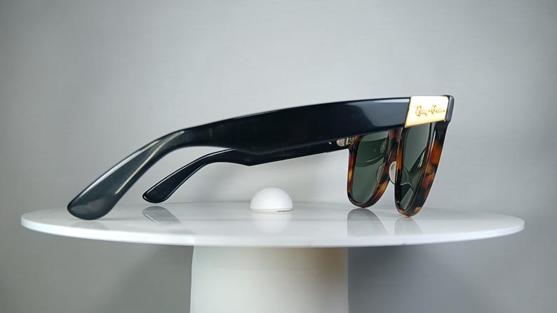 May include: Black sunglasses with a tortoiseshell pattern on the frames and green lenses. The sunglasses have a gold accent on the temples and are resting on a white surface.