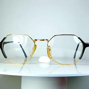 May include: A pair of eyeglasses with a gold-colored frame and black accents on the temples. The lenses are clear, and the glasses have a geometric, octagonal shape. The glasses are displayed on a white, round surface.