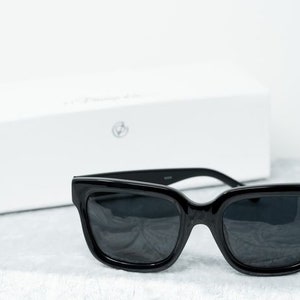 Puede incluir: Gafas de sol cuadradas negras con lentes oscuros. Las gafas de sol est&aacute;n sobre una superficie blanca.