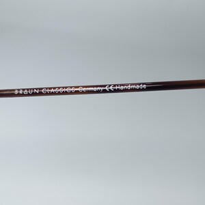 May include: Close-up of a brown and tan eyeglass arm. The text "BRAUN CLASSICS Germany CE Handmade" is printed in white along the arm. The arm has a pointed tip.