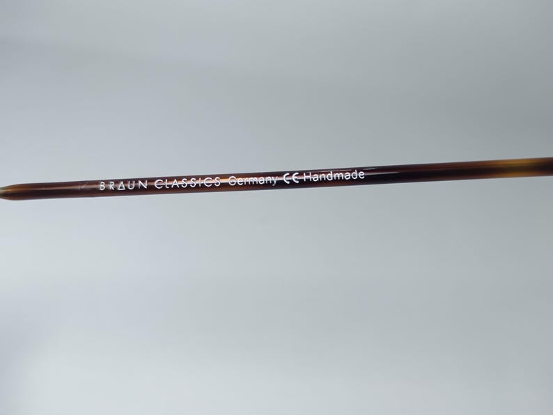 May include: Close-up of a brown and tan eyeglass arm. The text "BRAUN CLASSICS Germany CE Handmade" is printed in white along the arm. The arm has a pointed tip.