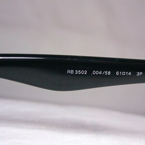 K&ouml;nnte beinhalten: Nahaufnahme des schwarzen B&uuml;gels einer Sonnenbrille mit dem aufgedruckten Text "RB 3502 .004/58 61□14 3P" auf der Innenseite.