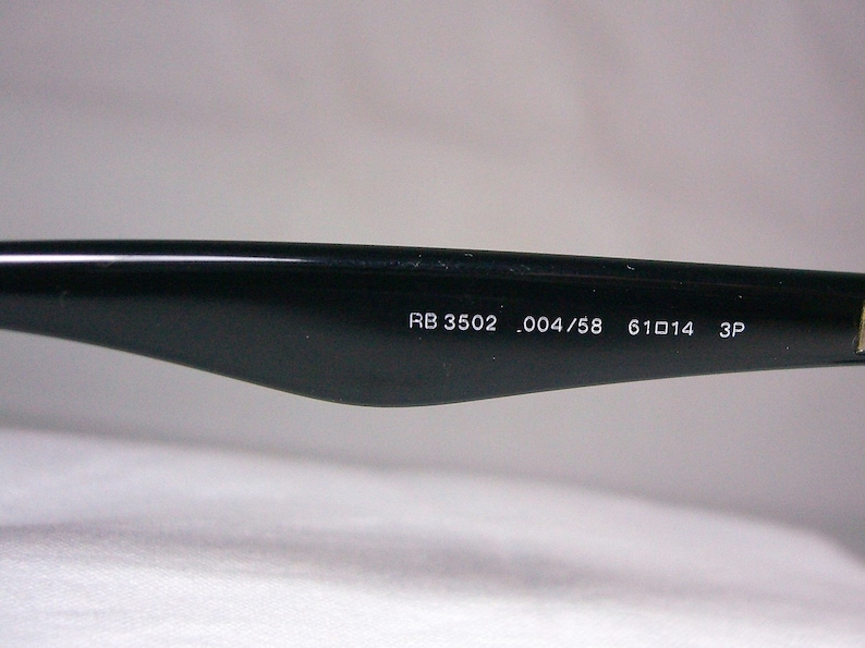 K&ouml;nnte beinhalten: Nahaufnahme des schwarzen B&uuml;gels einer Sonnenbrille mit dem aufgedruckten Text "RB 3502 .004/58 61□14 3P" auf der Innenseite.