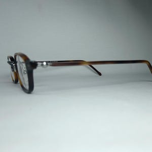 May include: Eyeglasses with a dark brown frame and tortoise shell arms. The lenses are a light amber color. The glasses are set against a white background.
